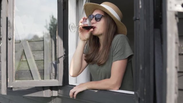 Beautiful woman in a hat is drinking red wine on a farm. Woman in the window with a glass of red wine