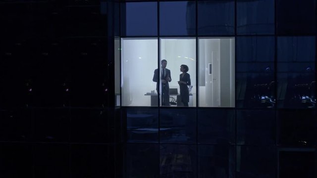Aerial Footage From Outside of the Skyscraper: Businessman and Businesswoman Talking while Standing in the Office Window. Retrieving Flying Shot of the Business District in the Evening