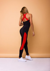 Obraz premium young sexy blonde girl in black costume with red stripes and white sneakers is standing from the back and looking down on the beige wall background, sport concept, free space