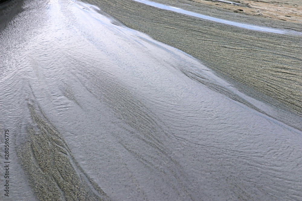 Wall mural Water flows over sand surface. Abstract gray natural background. Day view close-up of the textured stream with sky reflections.