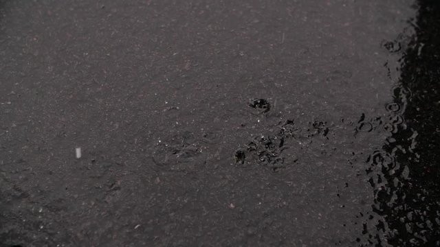 A slow motion shot of rain falling from the sky onto a dark grey rough concrete floor, the raindrops splatter with every drip. The floor is already soaked from the torrential downpour. 