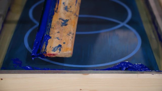 A close up of a screen printing frame being prepared, a man pours blue paint onto the screen and squeegee as he is preparing it to be used