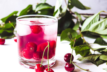 A glass of cherry drink with ice in a glass on a white wooden background. Fresh summer cocktail with cherries and ice cubes.