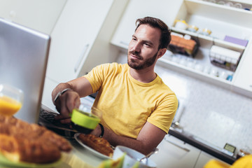 Busy man working at home, using laptop while having breakfast.