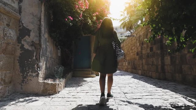 Young brunette with long hair walks down the street of Jaffa old city and spending her hand over flowers on tree, slow motion. Girl tourist in green dress walks around historic old town.