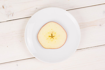 One slice of fresh apple red delicious on a small white ceramic plate flatlay on white wood
