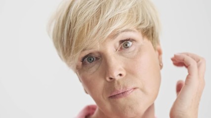 Cropped view of concentrated serious blonde old woman fixing her hair while looking at the camera over gray background isolated