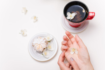 female hands holding spring jasmine flowers and sugar powdered cake with a red cup of green tea on the white brakfast table