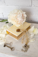 A beautiful white peony lies on a book with Golden pages. Near are delicate petals, vintage keys and embellishment on the neck. Vintage background in light white and beige.
