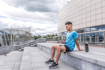 Athletic man summer city, sitting steps hands smartphone, online training social networks application Internet. Rest after exercise. Free space for text. Sportswear, shaker with protein water bottle.