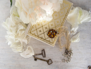 A beautiful white peony lies on a book with Golden pages. Near are delicate petals, vintage keys and embellishment on the neck. Vintage background in light white and beige.