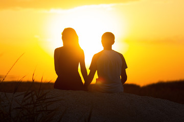 silhouette of a loving couple at sunset sitting on sand on the beach, the figure of a man and a...