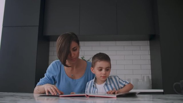 Young beautiful mother and son sitting at a table in a bright kitchen reading a book and looking at pictures poking a finger into a book and leafing through the pages.