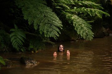A curly man swimming on his back on the silty brown water with face and feet raised up. Tropical river covered with big fern leaves