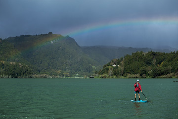 A man in shorts and red jacket floats standing on a sapsurf on the lake in cloudy weather with beautiful rainbow background. Furnas Lake, Azores