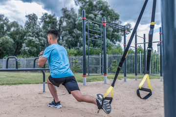 Young guy male athlete doing lunges look from back, squats city summer sports ground, webbing loops, leg muscle training, active lifestyle health sport concept, full-length sportswear, fitness watches