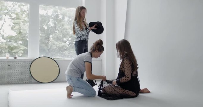 Backstage of the photo shoot: Photographer straightens clothes of a beautiful caucasian pregnant model while stylist cleans accessory in studio. Personal photoshoot. Studio photoshoot with professiona