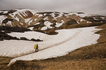 Hiker man is crossing the valley with snow and brown volcanic stones. Cloudy sky background. Landmannalaugar, Iceland