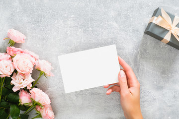 Hand holding blank paper card mockup on concrete stone desk with pink rose flowers and gift box. Flat lay, top view, copy space.