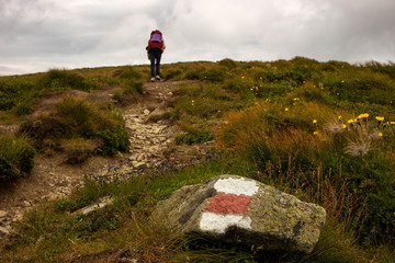Obraz premium Young girl with backpack trekking in mountain route. Route mark in foreground.