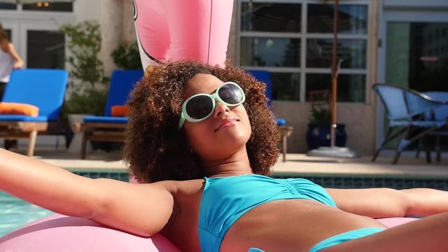 At the swimming pool, a beautiful mixed-race woman enjoys the warmth of the sun as she relaxes on a pink flamingo floatie.