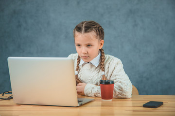 The little blond girl with braids is concentrated on the grey laptop, watching to a screen and having a cup of hot coffee near her.