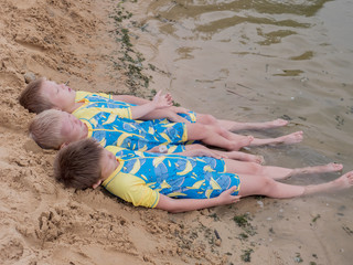 Funny kids in bathing suits sit together on sand with their legs in water. boys are happy on holiday in village together. Summer day, river, swimming in the water. Close-up legs and feet