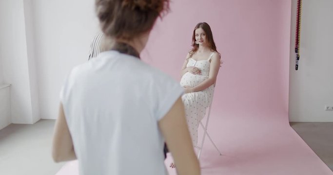Backstage of the photo shoot: Stylist straighten hair of a beautiful caucasian pregnant model in a moment photographer starts taking photos with professional camera. 4K RAW graded footage 24fps