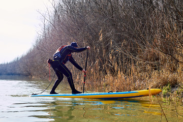Man on stand up paddle boarding (SUP) paddling along the calm autumn Danube river against a background of brown autumn trees at the shore. Concept of water tourism, healthy lifestyle and recreation