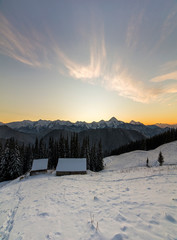 Old wooden house, hut and barn in deep snow on mountain valley, spruce forest, woody hills on clear blue sky at sunrise copy space background. Mountain winter panorama landscape.