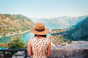 Rear view of young woman in dress and hat, who looking from above to the Kotor city and Boka bay, Montenegro - obrazy, fototapety, plakaty
