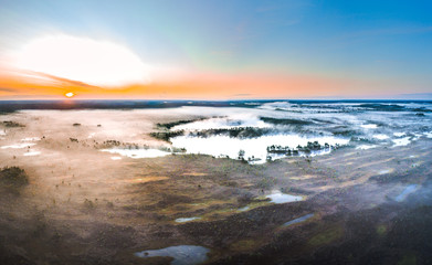 Warmly colored sunrise over a foggy swamp. Aerial view of stunning landscape at peat bog at Kemeri national park in Latvia. Wooden trail leading along the lake surrounded by pounds and forest. 