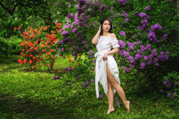 Barefoot girl in a white dress walks in the flowered garden. Lilac bushes
