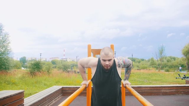 A strong man with tattoos pulling up on parallel bars on a sports ground