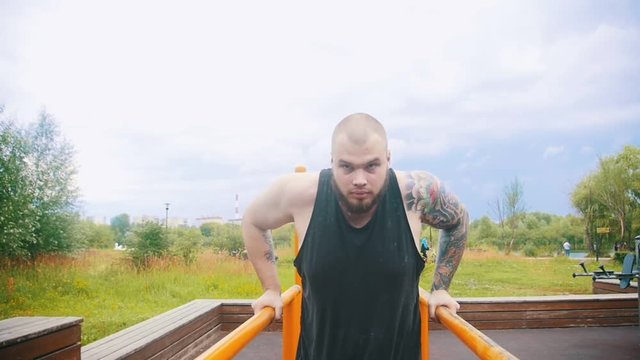A strong bearded man with tattoos pulling up on parallel bars on a sports ground