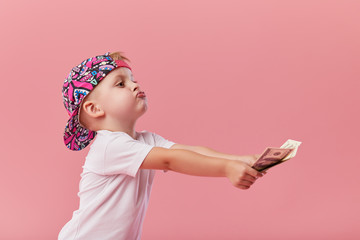 Portrait of a cheerful little toddler boy in cap holding a dollars money over pink background