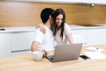 Young embracing couple is using laptop computer sitting on kitchen at home