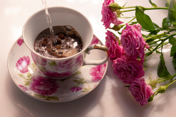 Hot water is poured into a cup for making morning coffee. A cup of coffee with pink decor and flowers of roses on the table in the morning sun.