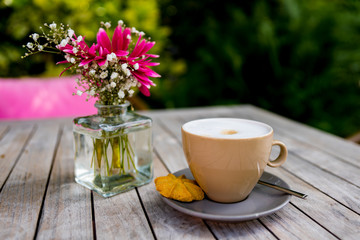 Cup of cappuccion on wooden table and a small bottle with pink gerbera and white twig._