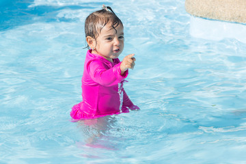 Fototapeta na wymiar Baby girl of two years old stands playing in a swimming pool. She is happy and playful with her experience.