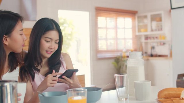 Asian Lesbian lgbtq women couple have breakfast at home, Young Asia lover girls happy using mobile phone check news while drink juice, cornflakes cereal and milk in kitchen in the morning concept.