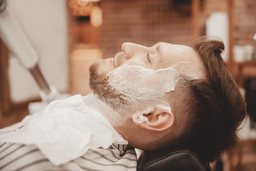 Barber shaves beard of man in barbershop with razor