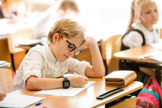 Happy cute clever boy is sitting at a desk in a glasses with raising hand. Child is ready to answer with a blackboard on a background. Ready for school. Back to school