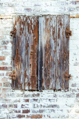 Closed brown red peeling wooden window with rusted metal hinges on a grungy white brick wall of an old abandoned derelict barn weathered by exposure to the elements and lack of maintenance.