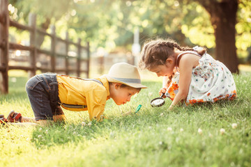 Cute adorable Caucasian girl and boy looking at plants grass in park through magnifying glass. Children friends siblings with loupe studying learning nature outside. Child education concept. - obrazy, fototapety, plakaty