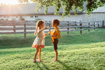 Funny Caucasian children girl and boy blowing soap bubbles in park at summer sunset. Real authentic happy childhood moment of friendship. Lifestyle activity for friends siblings.