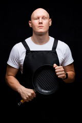 Handsome bald muscular young man in an apron holding a frying pan with emotional facial expression. Isolated on black background. Chef or husband concept.