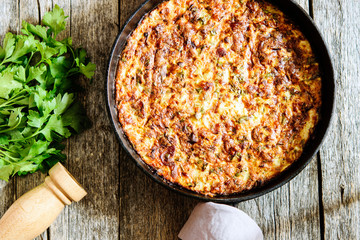 Zucchini Casserole with Bacon and Cheese in cast iron pan and parsley bunch and pepper mill on vintage wooden table. Bright sunlight. Selective focus
