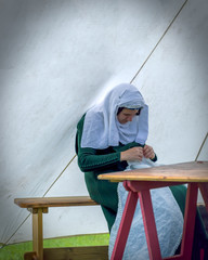 Woman in authentic medieval costume sat on a bench and table, sewing in a tent. The encampment at Cardiff Joust 2019.