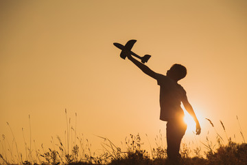 Black silhouette of young caucasian kid isolated on sunny golden sunset sky background. Boy playing toy plane outside on grassy summer hill. Setting big goals and dreaming about happy future concept. - Powered by Adobe
